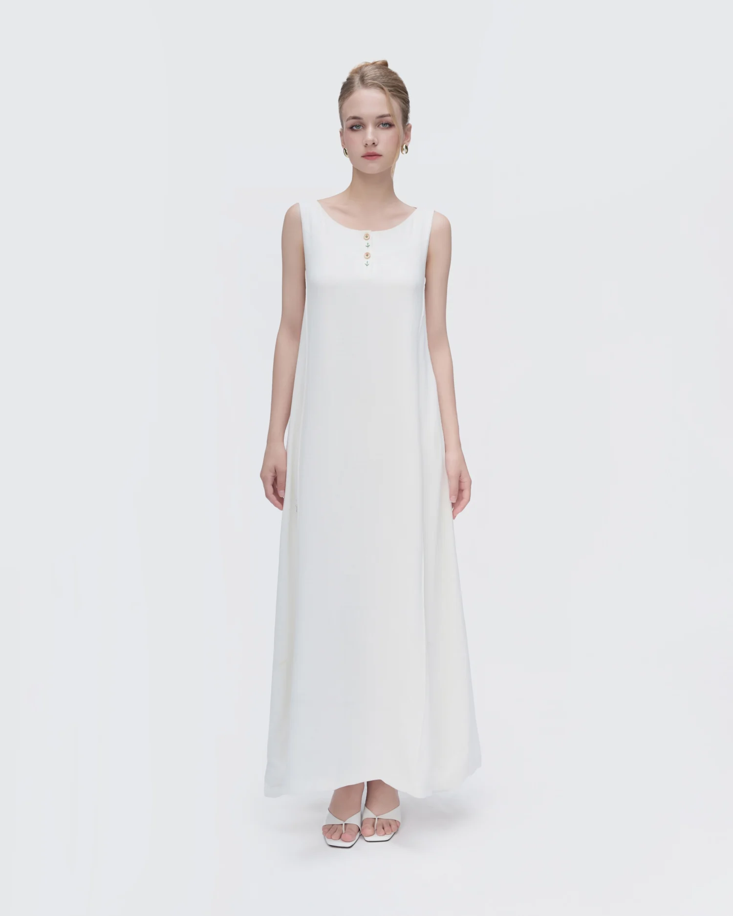 Woman in elegant white sleeveless maxi dress with button detail, wearing hoop earrings and sandals against a neutral background.