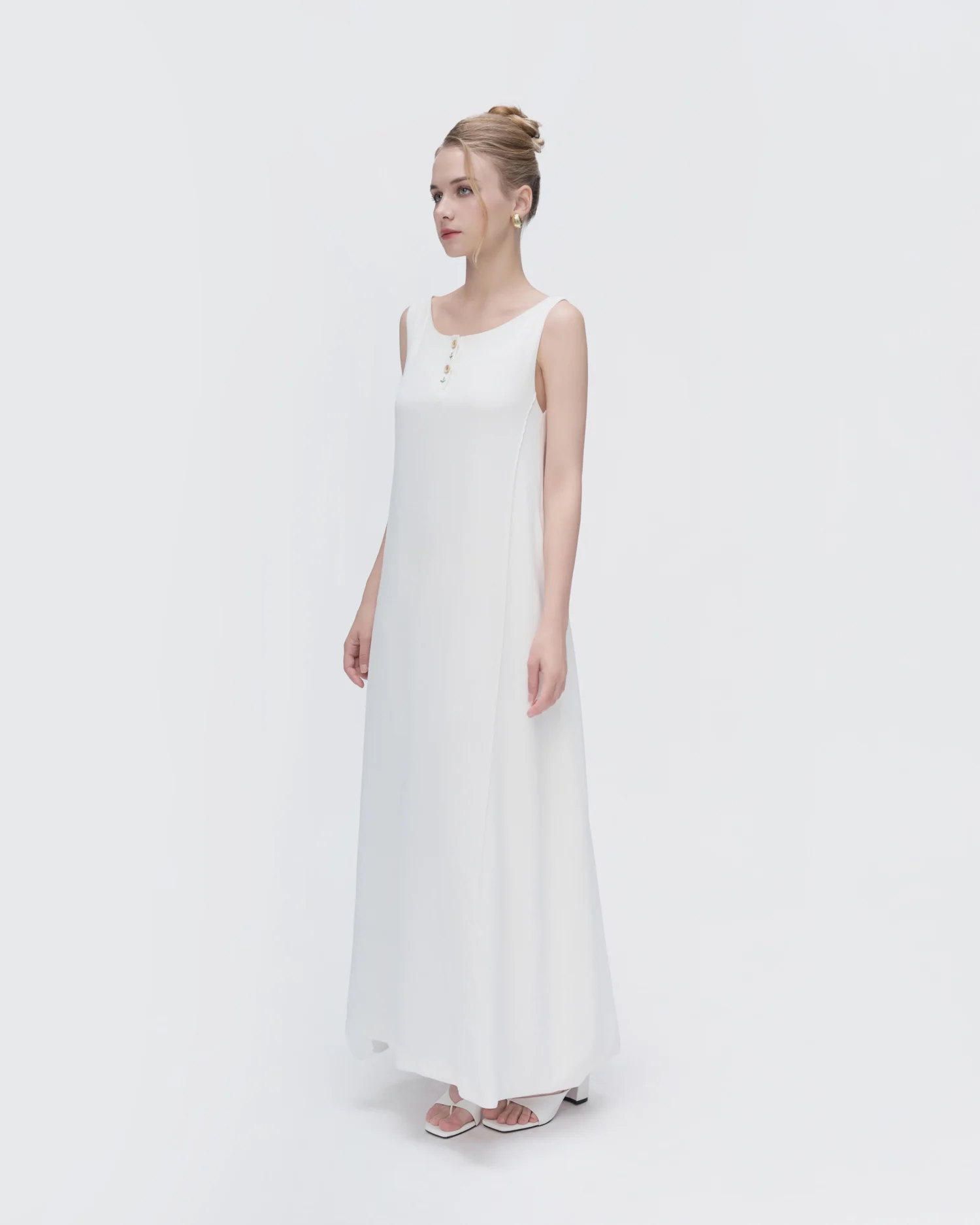 Elegant woman in a long white dress and high sandals, standing against a plain background.