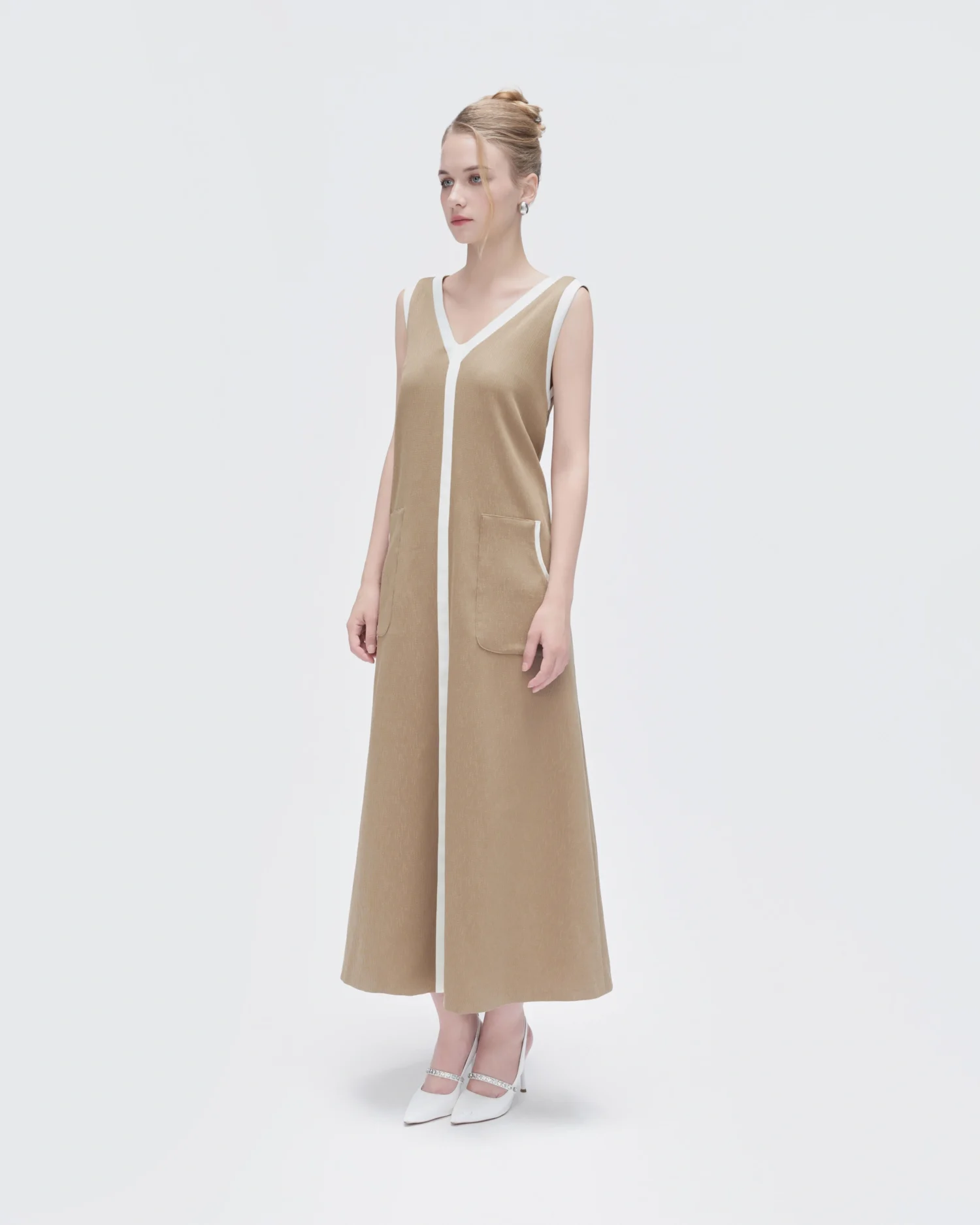 Woman in beige sleeveless dress with white trim, paired with white heels, standing against a plain background.