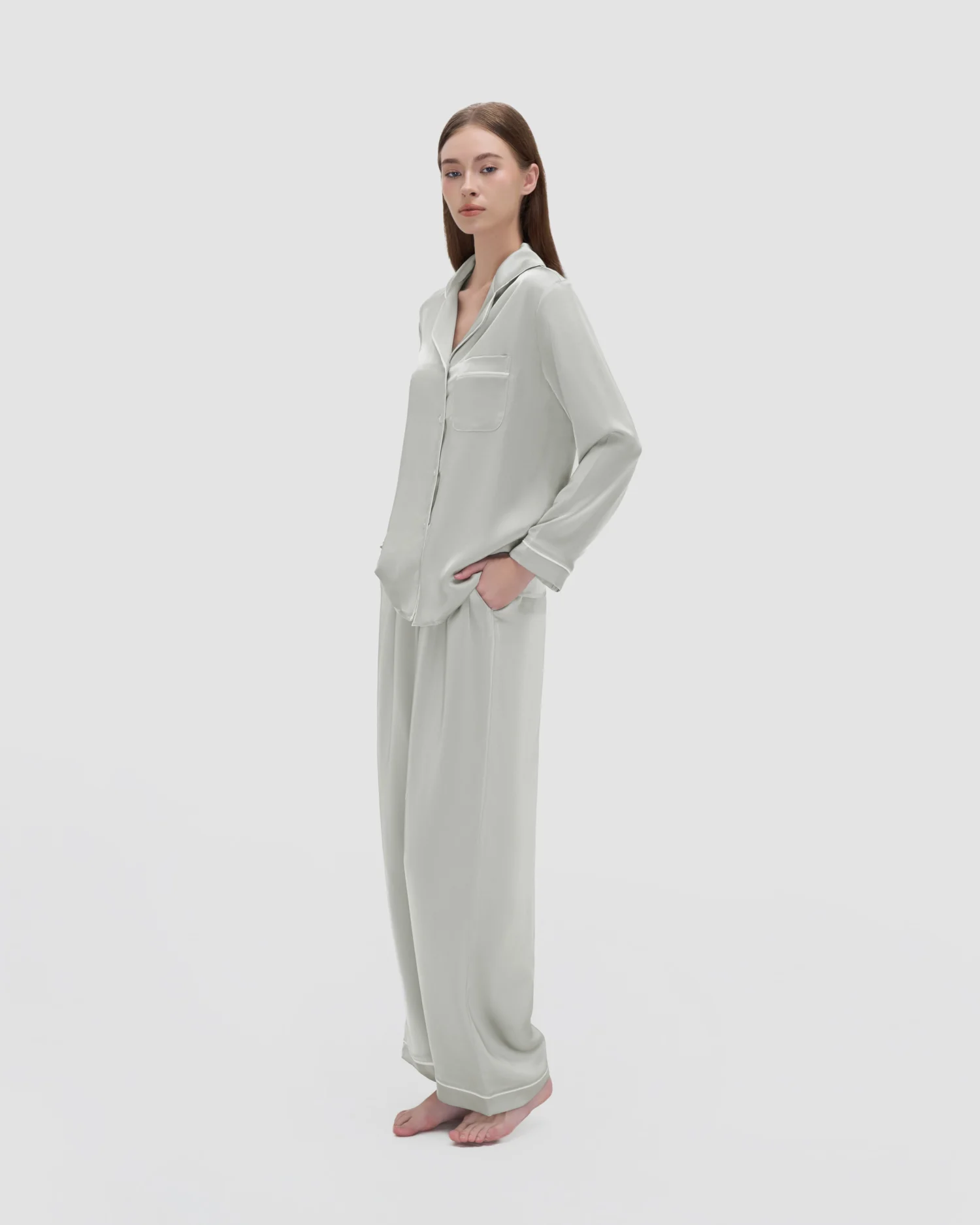 Woman in light grey silk pajamas standing barefoot against a plain background.