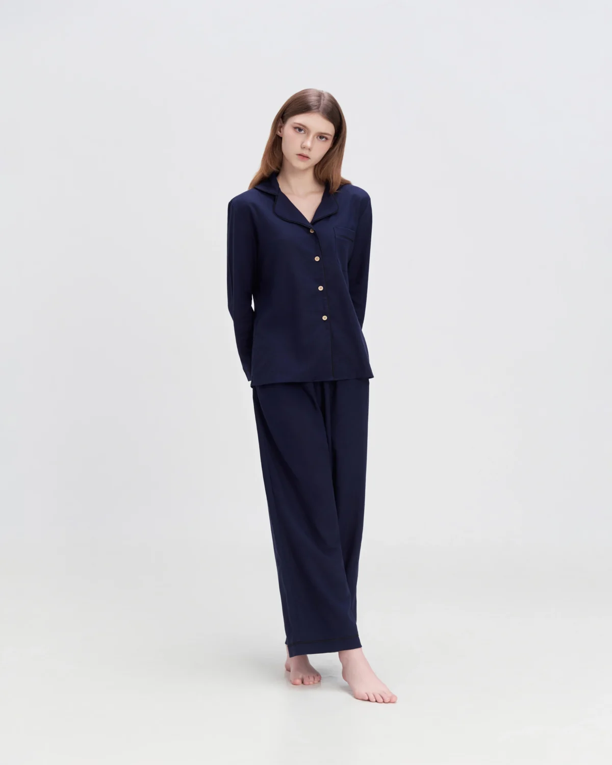 Young woman in navy blue button-up pajamas stands barefoot against a plain white background.