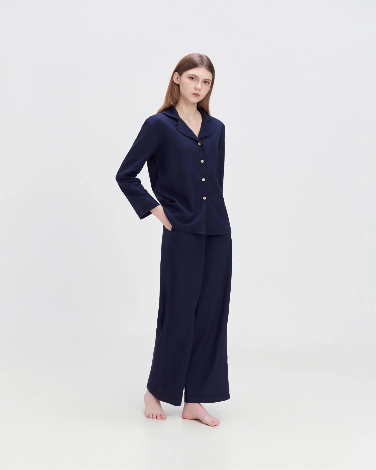 Woman in navy silk button-up pajama set standing against a white background, hands in pockets, casual and elegant.