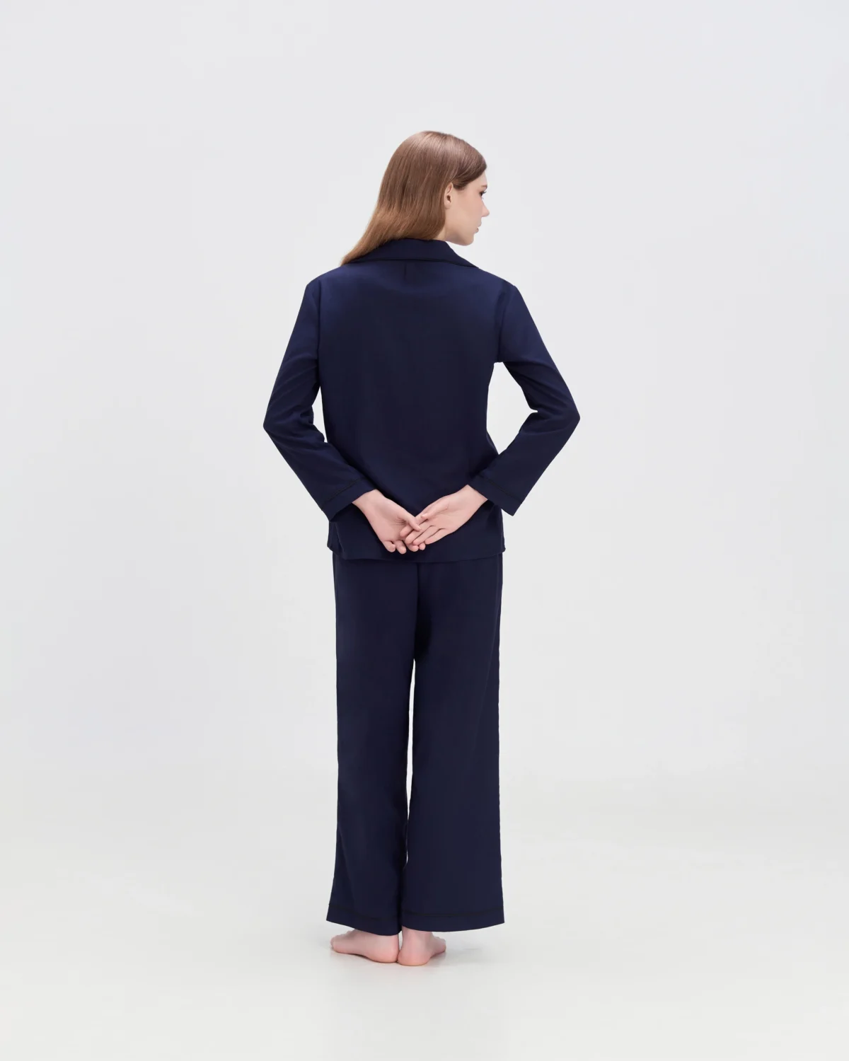 Woman in navy loungewear from behind with hands clasped, standing barefoot on a light background.