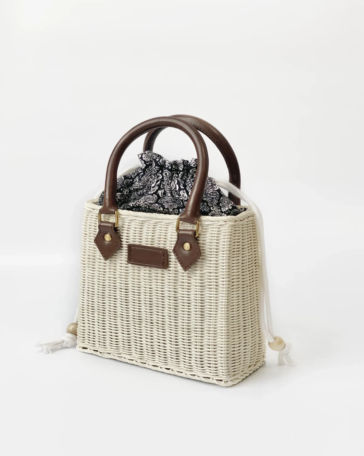 Woven handbag with dark handles and paisley fabric peeking out, on a white background. Ideal for stylish summer outings.