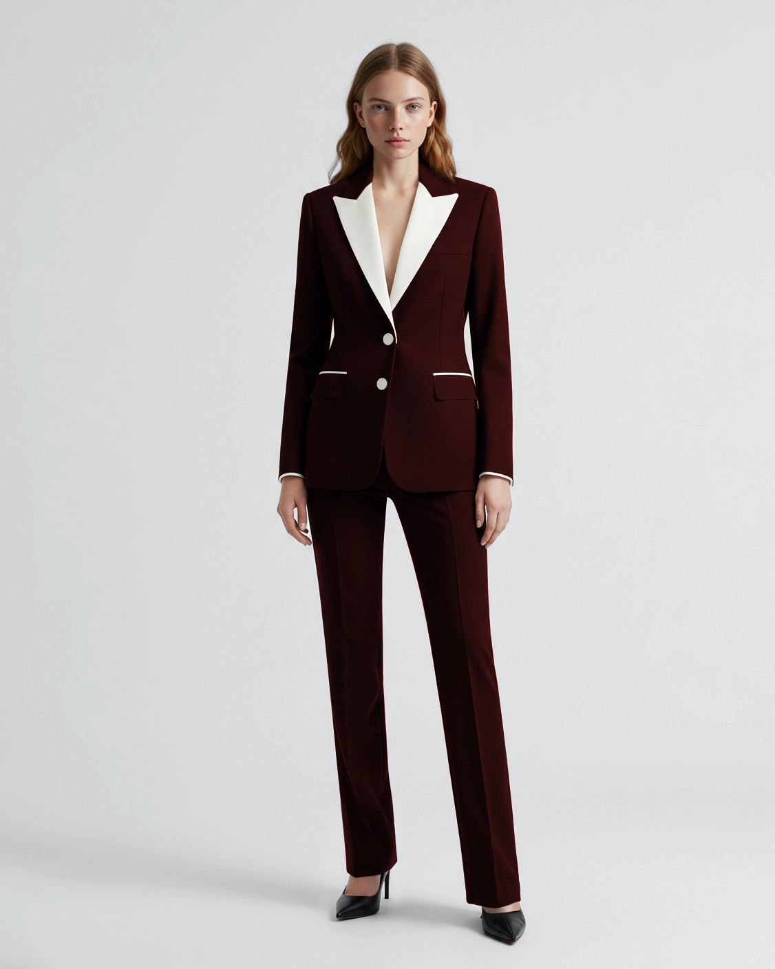 Woman in maroon blazer with white lapels, matching pants, and black heels stands confidently on a white background.