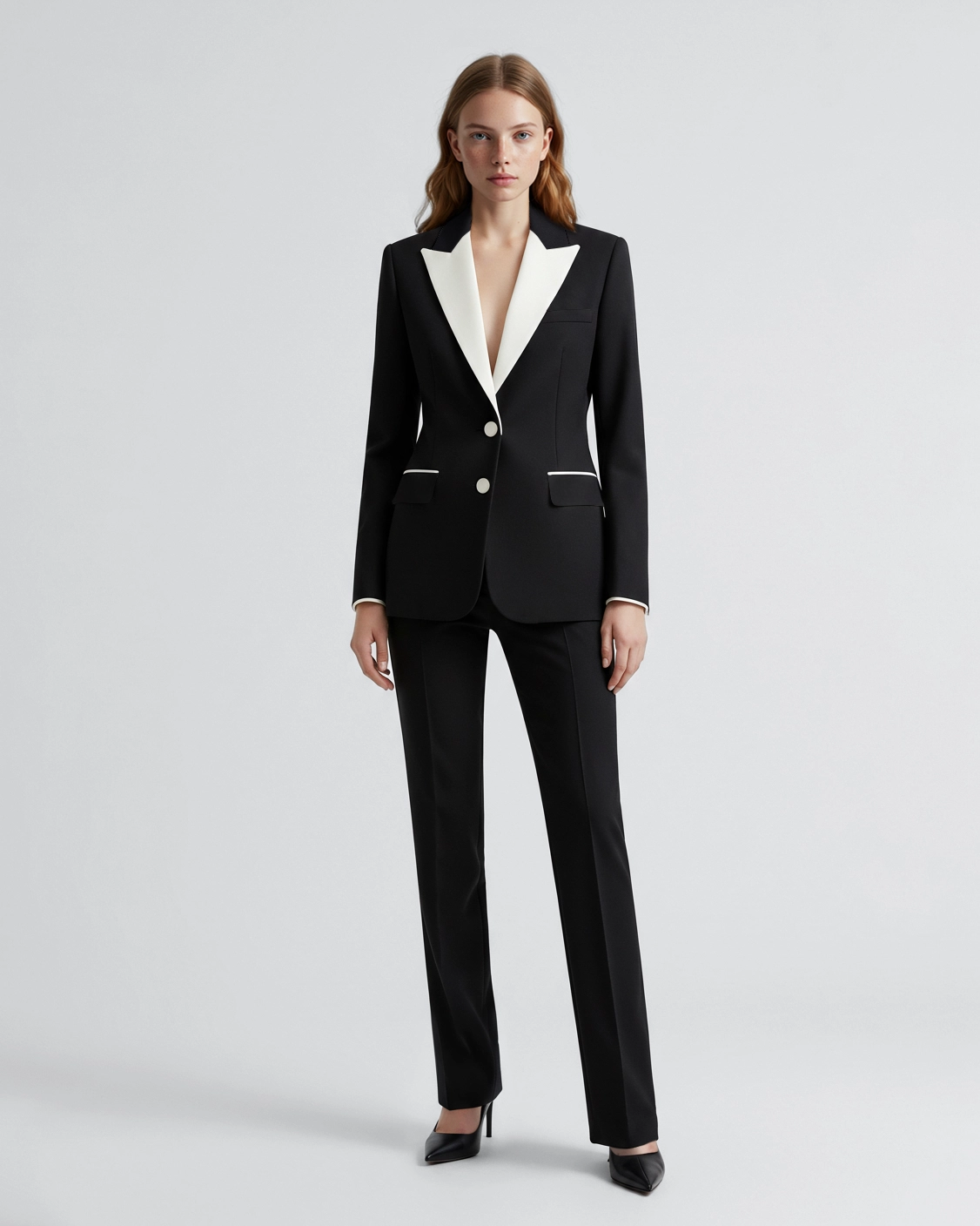 Woman in a stylish black tuxedo suit with white accents, standing confidently on grey background.