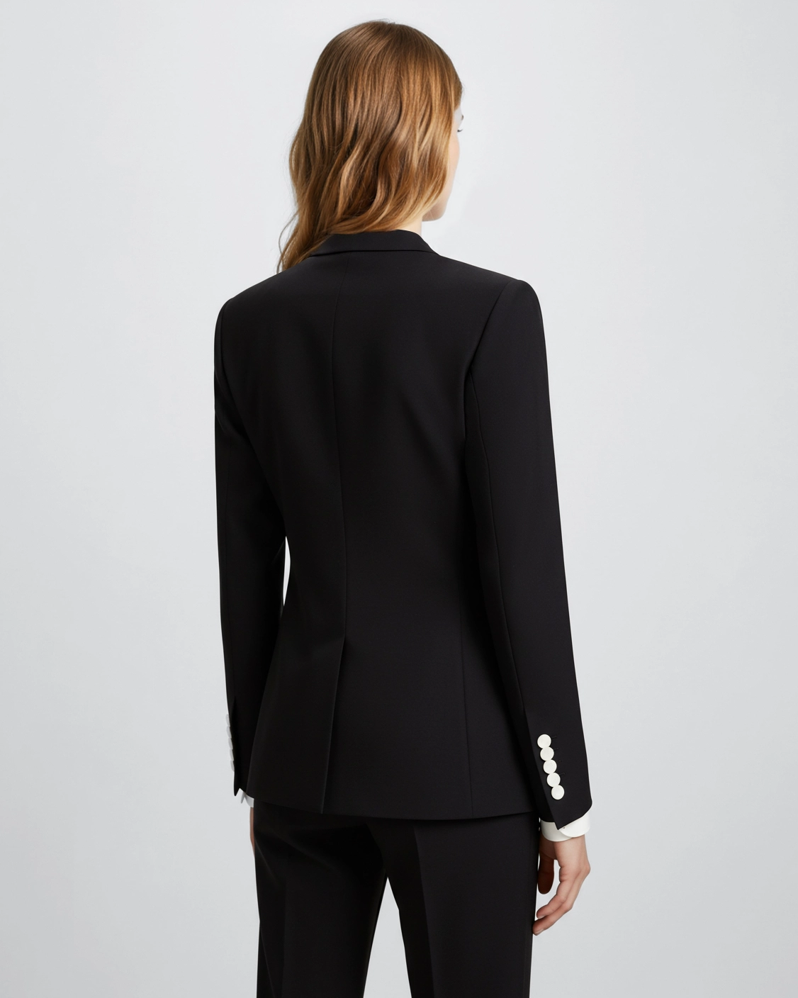 Rear view of a woman in a black suit with buttoned sleeves, standing against a plain white background.