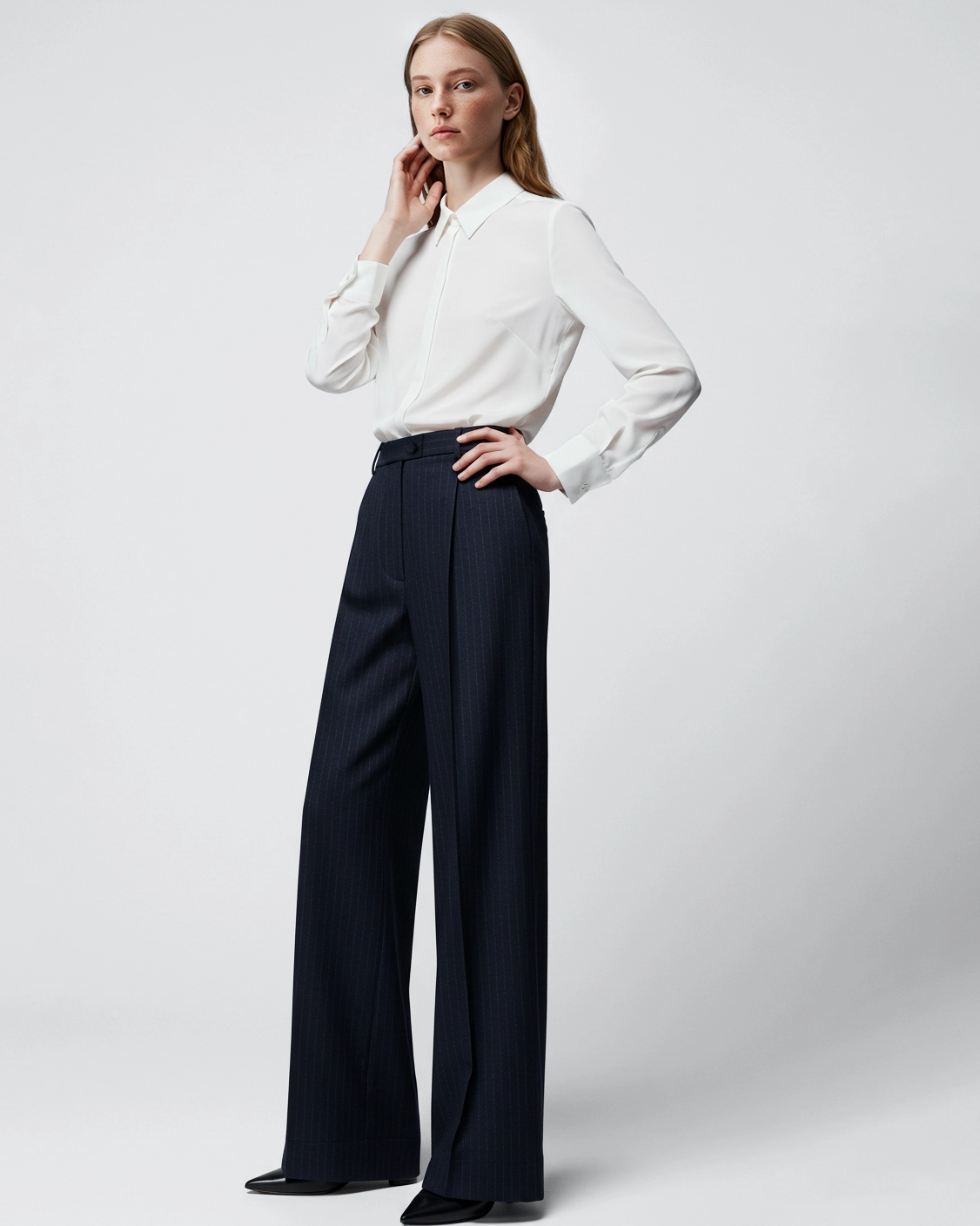 Woman in white blouse and navy wide-leg pants posing against a plain background.