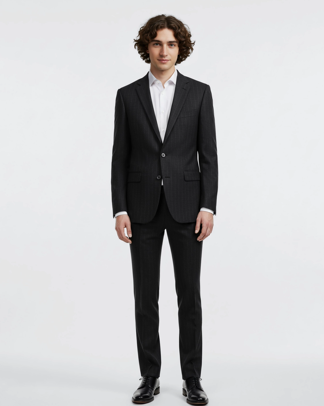 Man in a black suit and white shirt standing against white background, showcasing formal wear style.