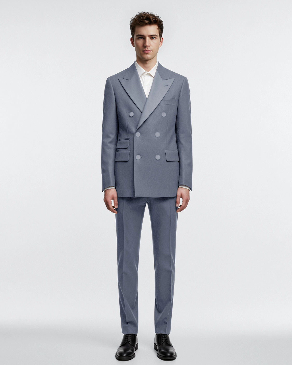 Man in a stylish gray double-breasted suit and black shoes, standing against a white background. Formal fashion.