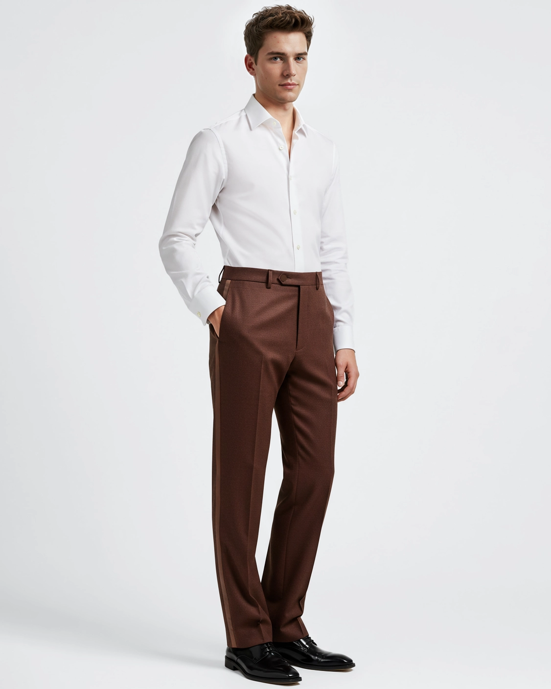 Man in a white dress shirt and brown pants, hands in pockets, standing against a white background.