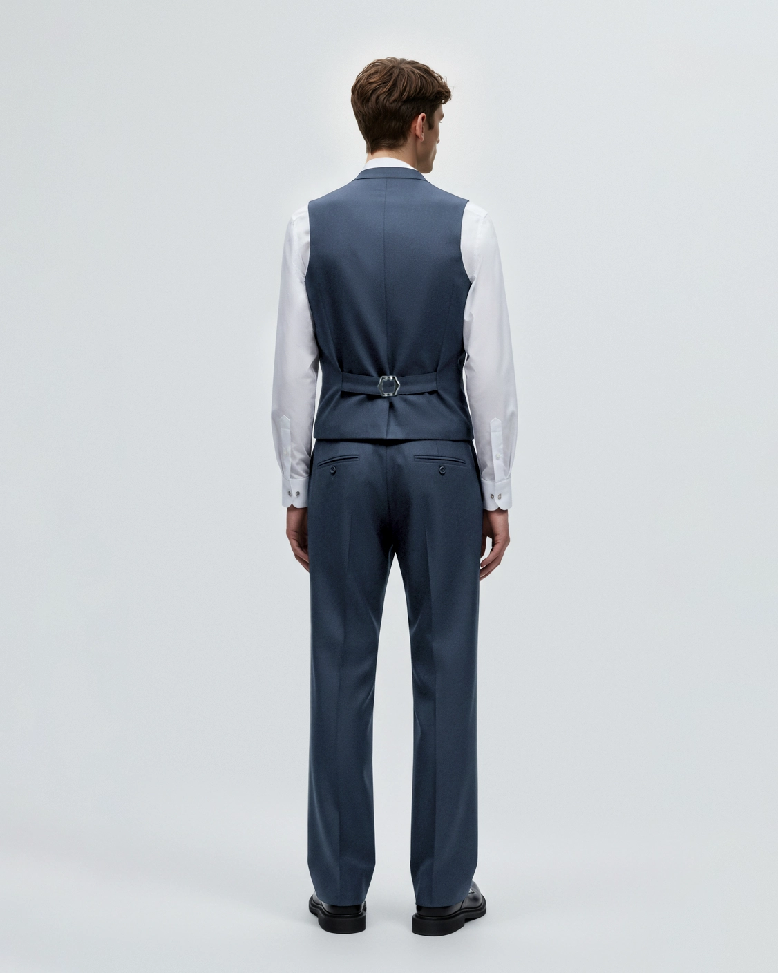 Man in blue tailored suit with vest and white shirt, viewed from behind against a plain background.