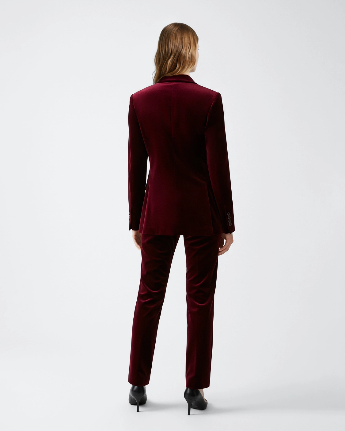 Back view of person in elegant burgundy velvet suit and heels on white background.
