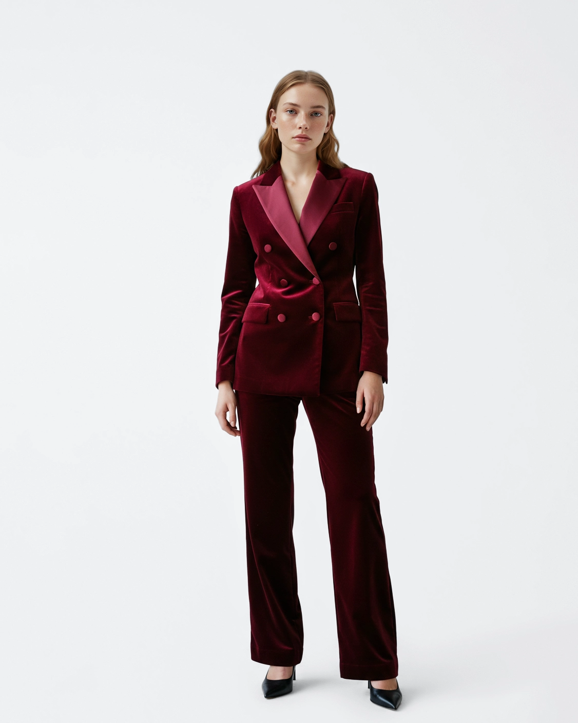 Woman in stylish maroon velvet suit with black heels, standing confidently against a white background.