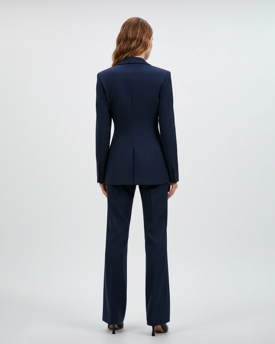 Woman in a navy suit, viewed from the back, stands against a plain backdrop, showcasing tailored fit.