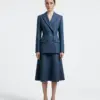 Woman in blue tailored suit and skirt standing against plain background.