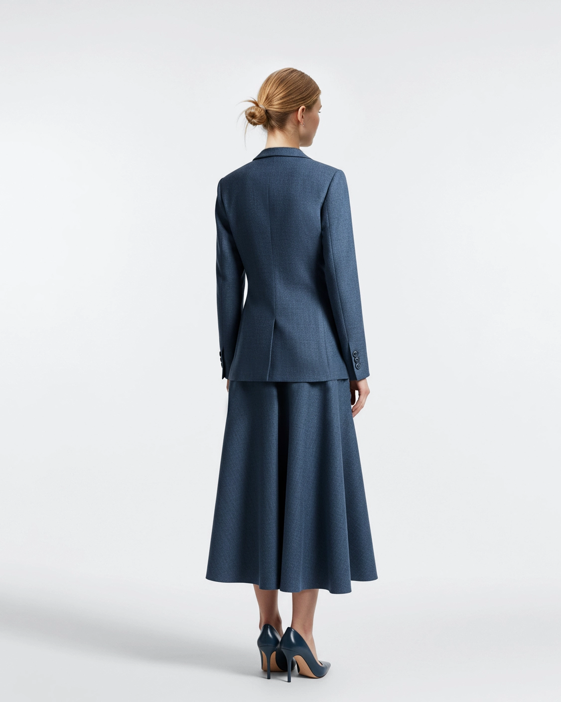 Woman in elegant blue suit and skirt, standing with back to the camera, in stylish high heels against white background.