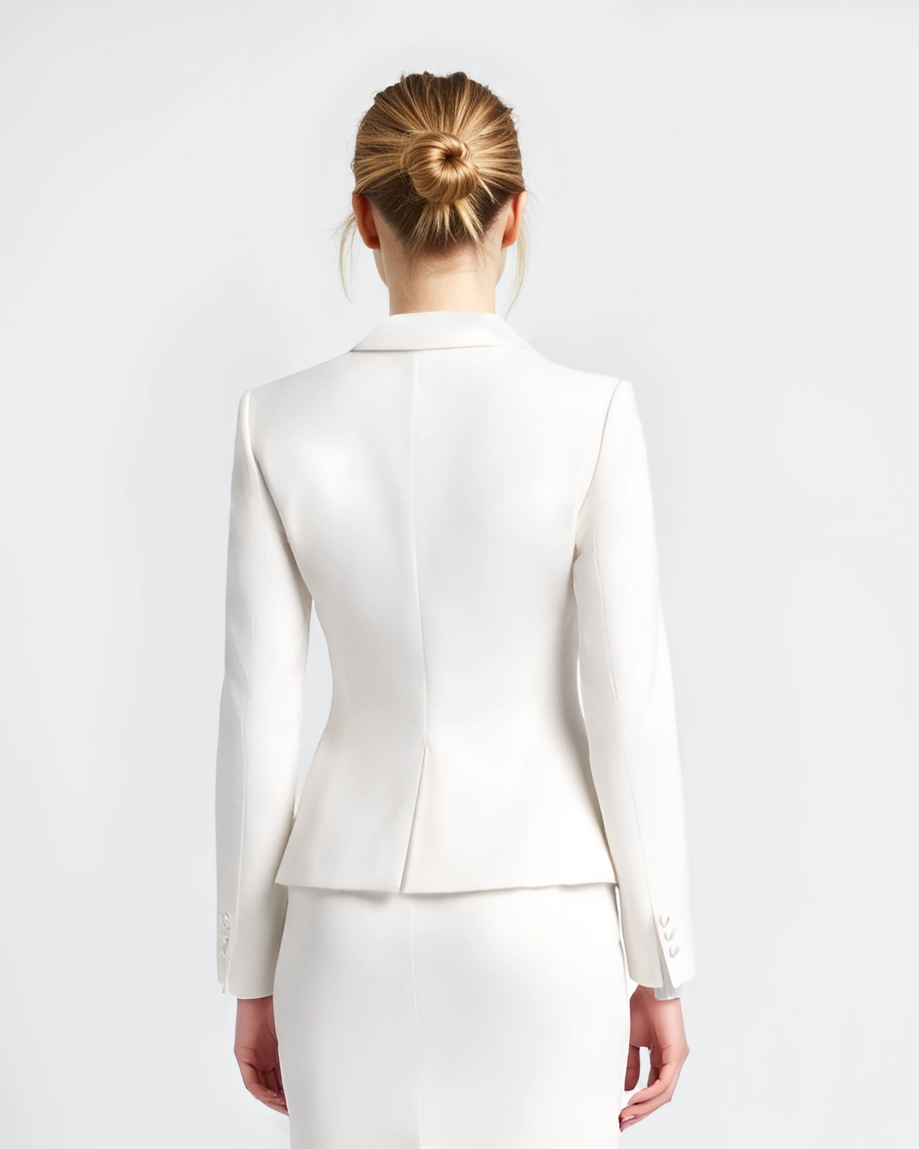 Back view of a woman in a white blazer and skirt with hair in a bun, standing against a neutral background.