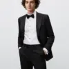 Young man in a black tuxedo with bow tie, smiling confidently with hands in pockets, against a neutral background.