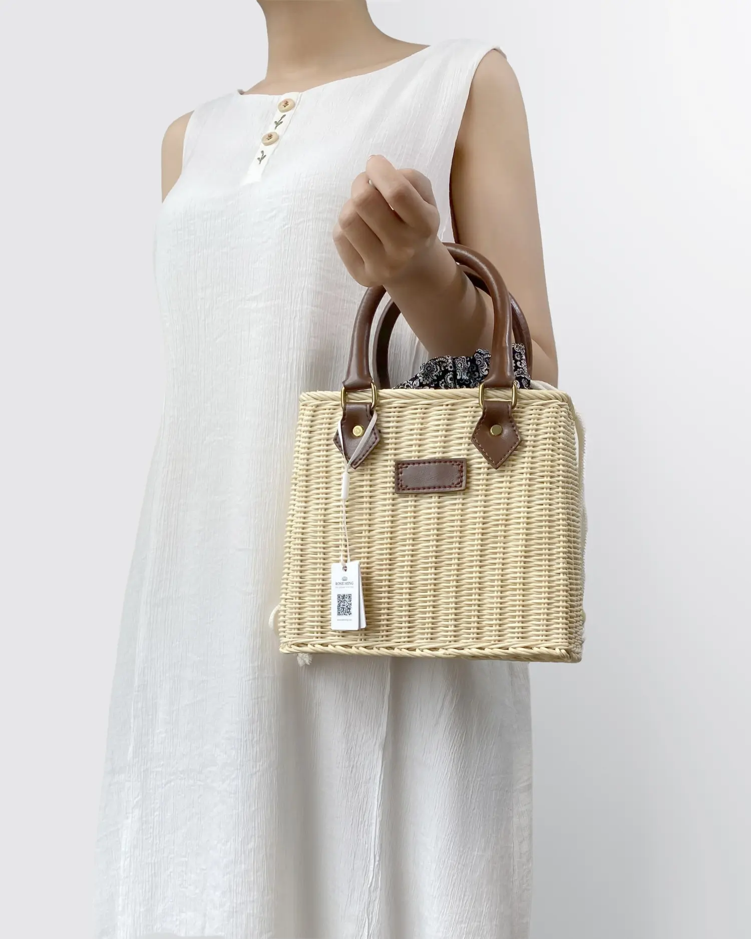 Woman in white dress holding a stylish woven handbag with leather handles and a tag.
