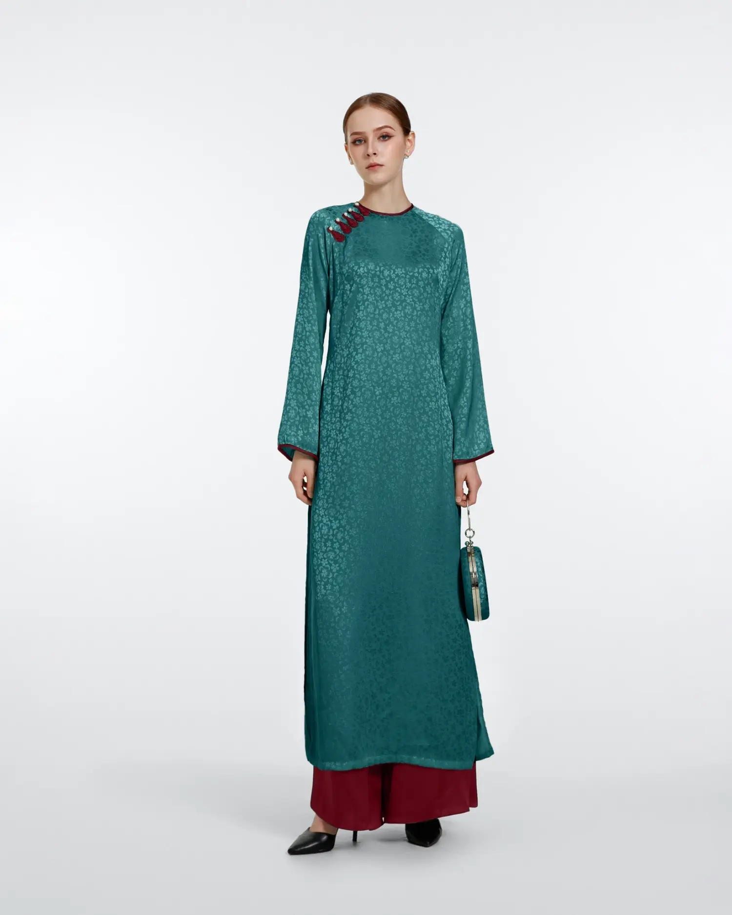 Woman in a teal traditional dress holding a matching handbag, wearing red pants and black heels on a white background.