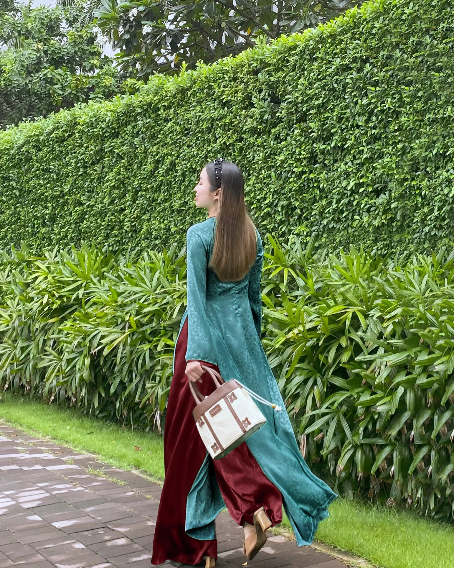 Woman in elegent green ao dai and red paint walking by lush green hedge, carrying a raffia handbag.