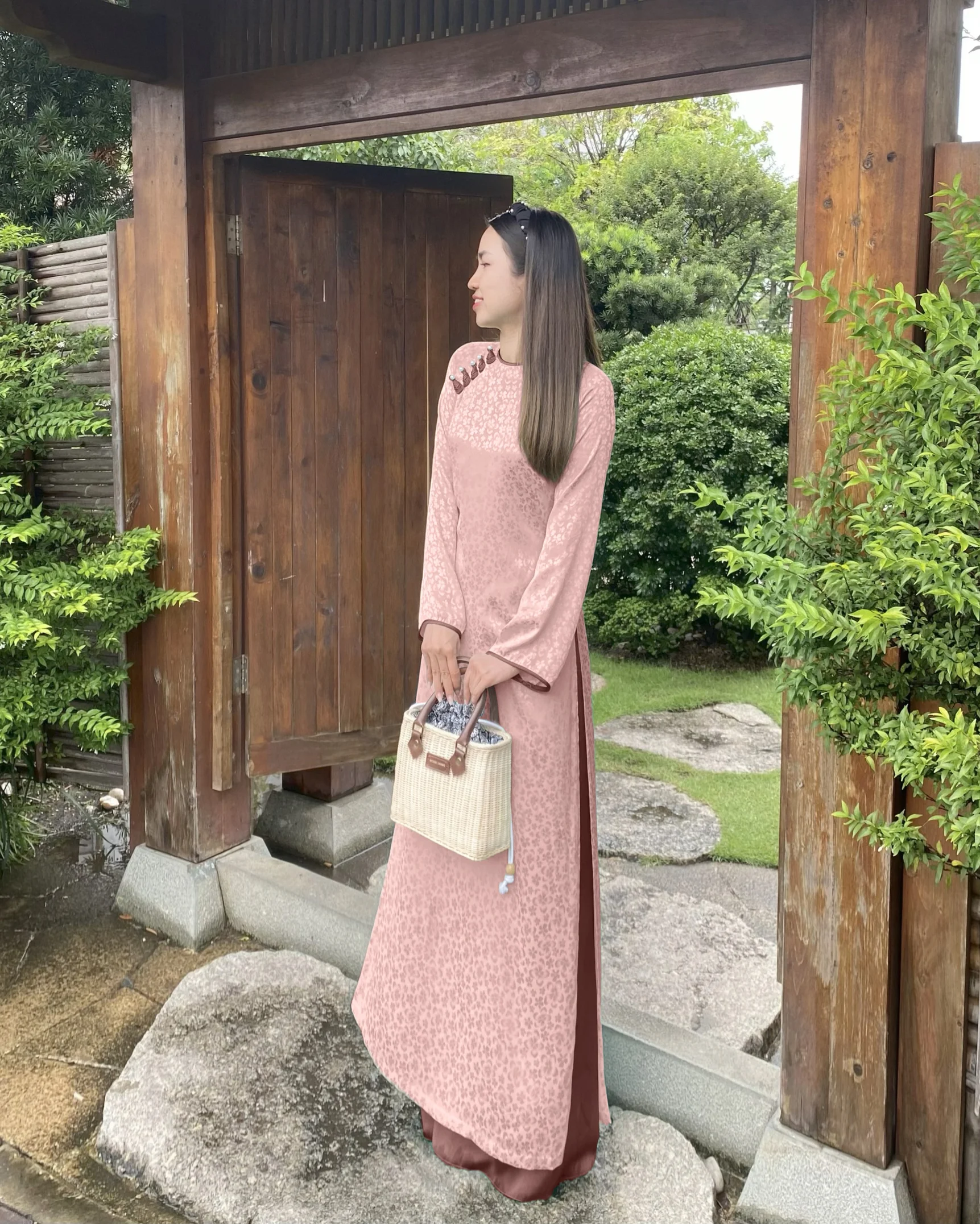 Woman in pink traditional dress stands by a wooden gate, holding a woven bag, surrounded by greenery.