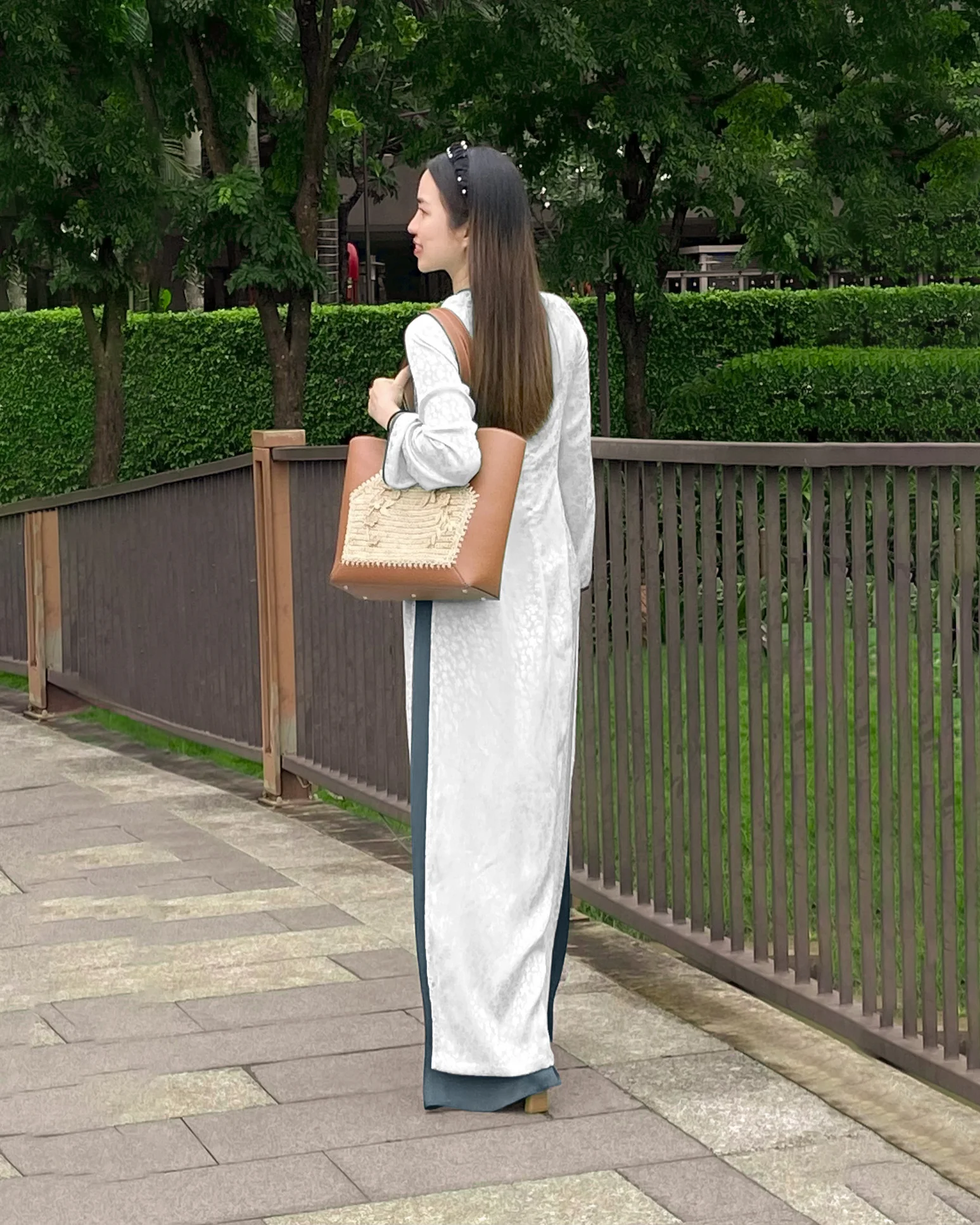 Woman in elegant ao dai with brown crocheted raffia bag, walking on a pathway surrounded by green trees.