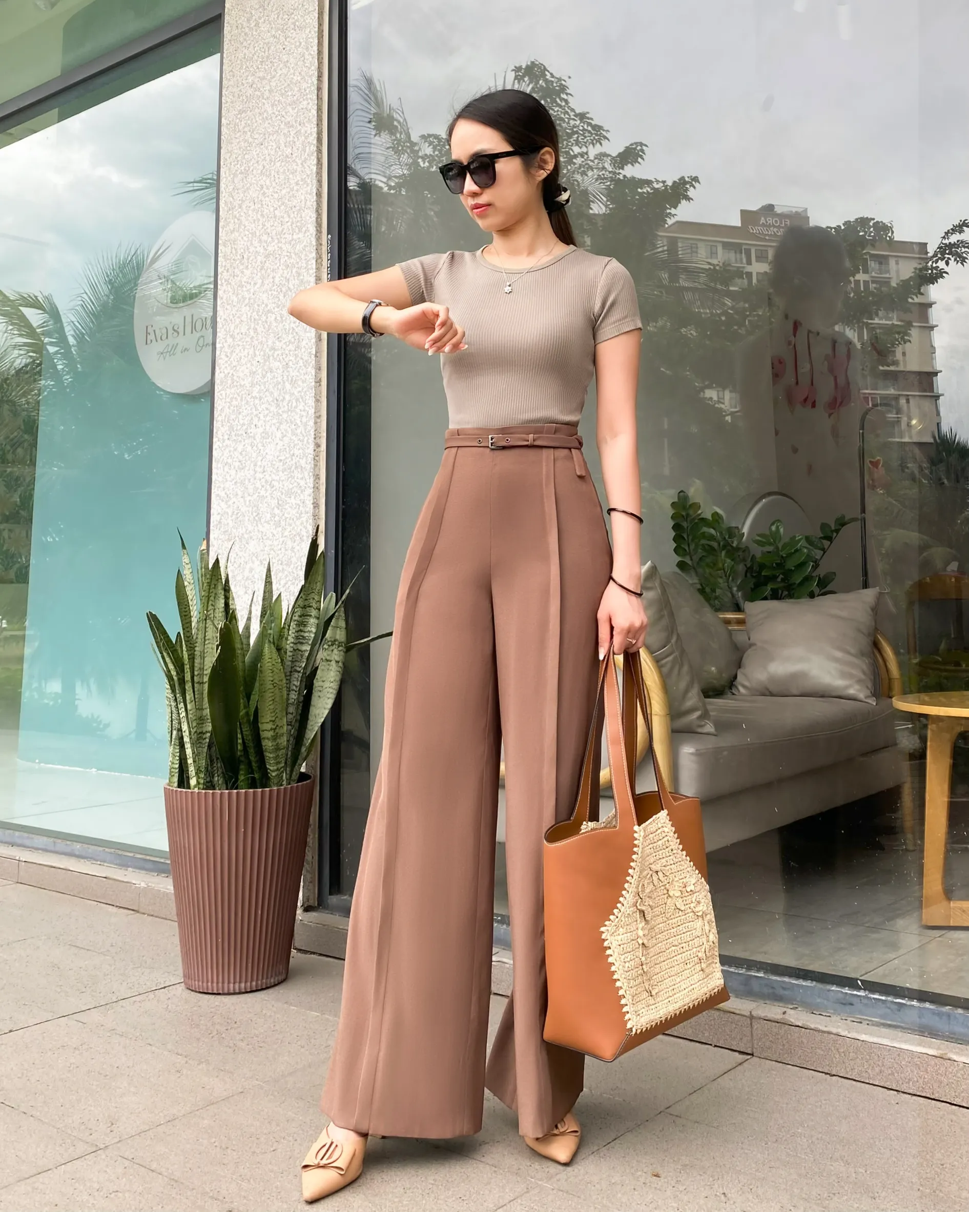 Fashionable woman in brown tailored trousers, sunglasses, checking time, holding crocheted raffia bag in front of glass storefront.