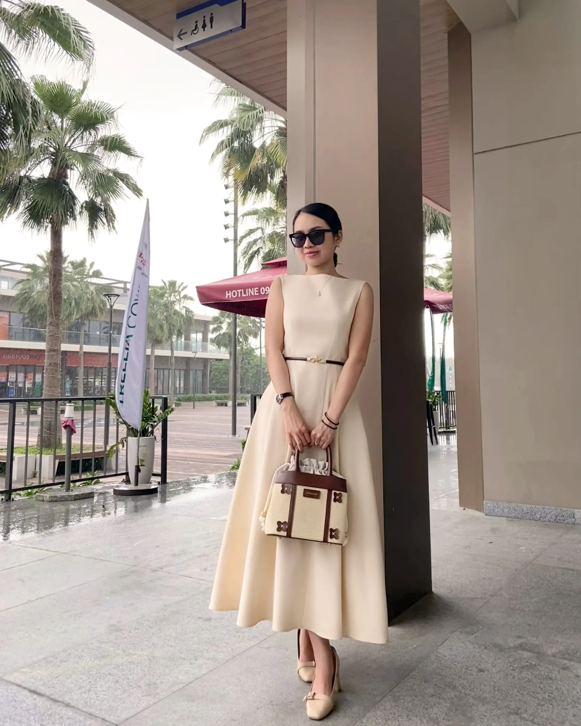 Woman in elegant beige dress with handbag stands under palm trees by a modern building.