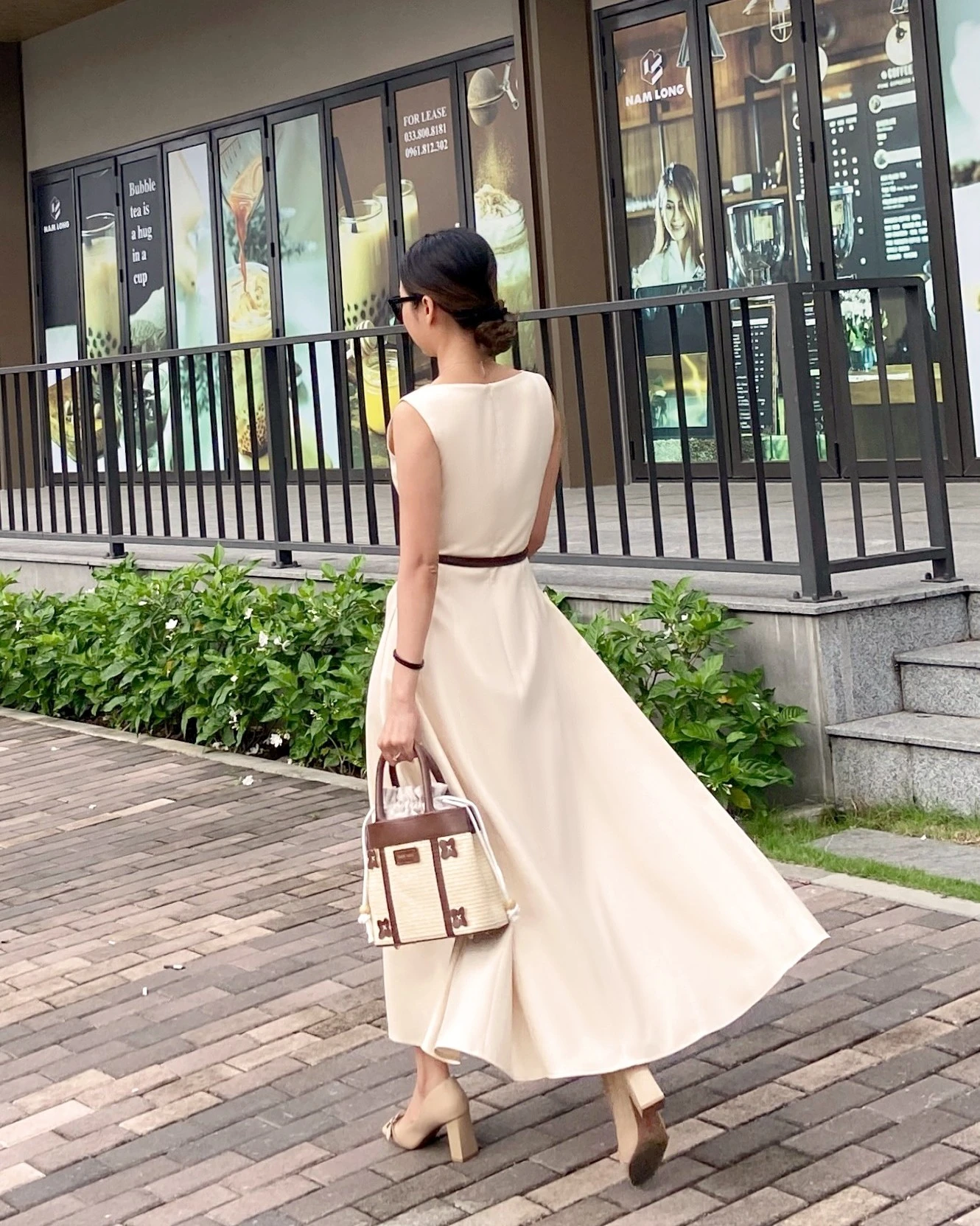 Woman in elegant beige dress and heels walking with a handbag by shop windows.