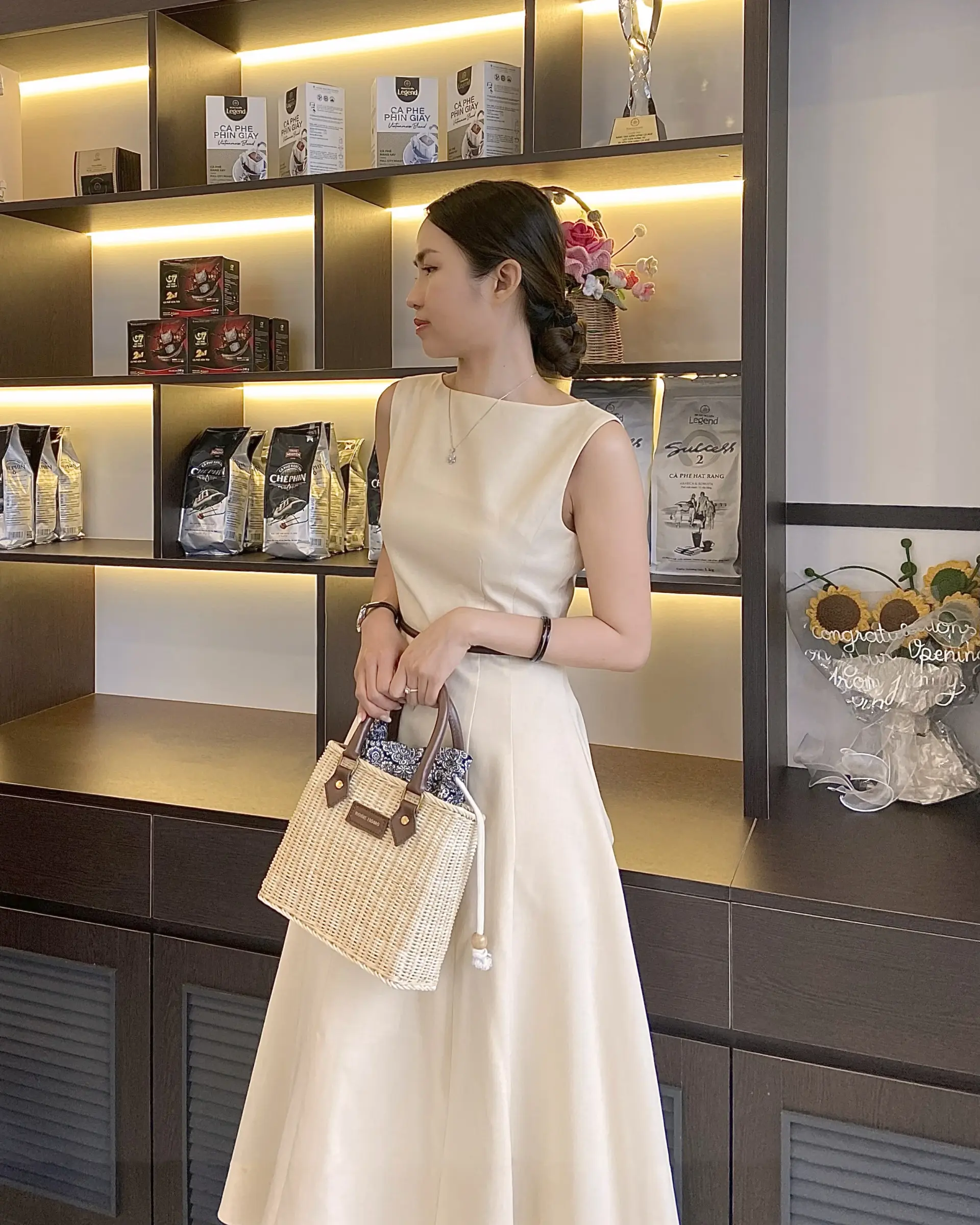 Woman in elegant dress holding a woven handbag, standing near illuminated shelves with coffee products and decorations.