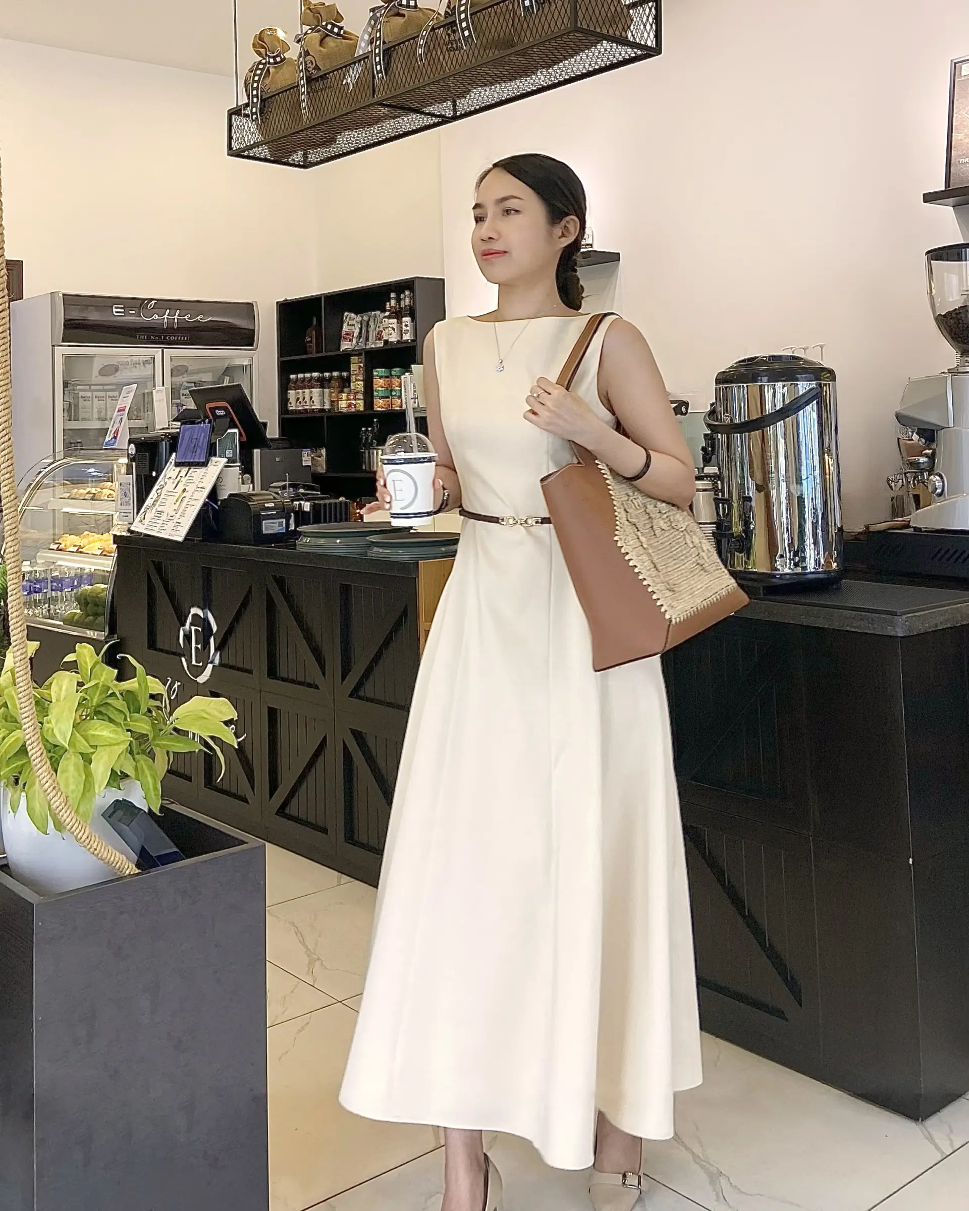 Woman in elegant ivory dress holding coffee, standing in a chic café, brown crocheted raffia tote bag on shoulder.