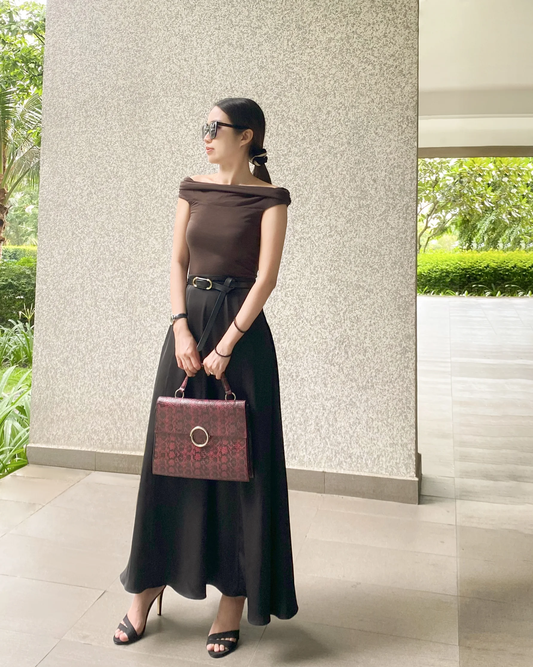 Stylish woman in brown top, black tailored flare skirt, holding a red handbag, wearing sunglasses, standing outdoors.