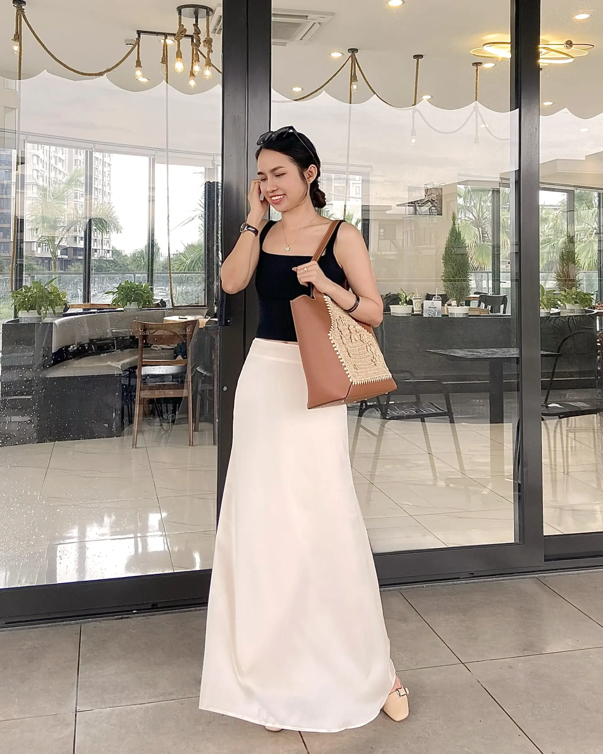 Woman in stylish black top and creamy silk skirt outside a café, carrying a brown chrocheted raffia tote bag, smiling and on the phone.