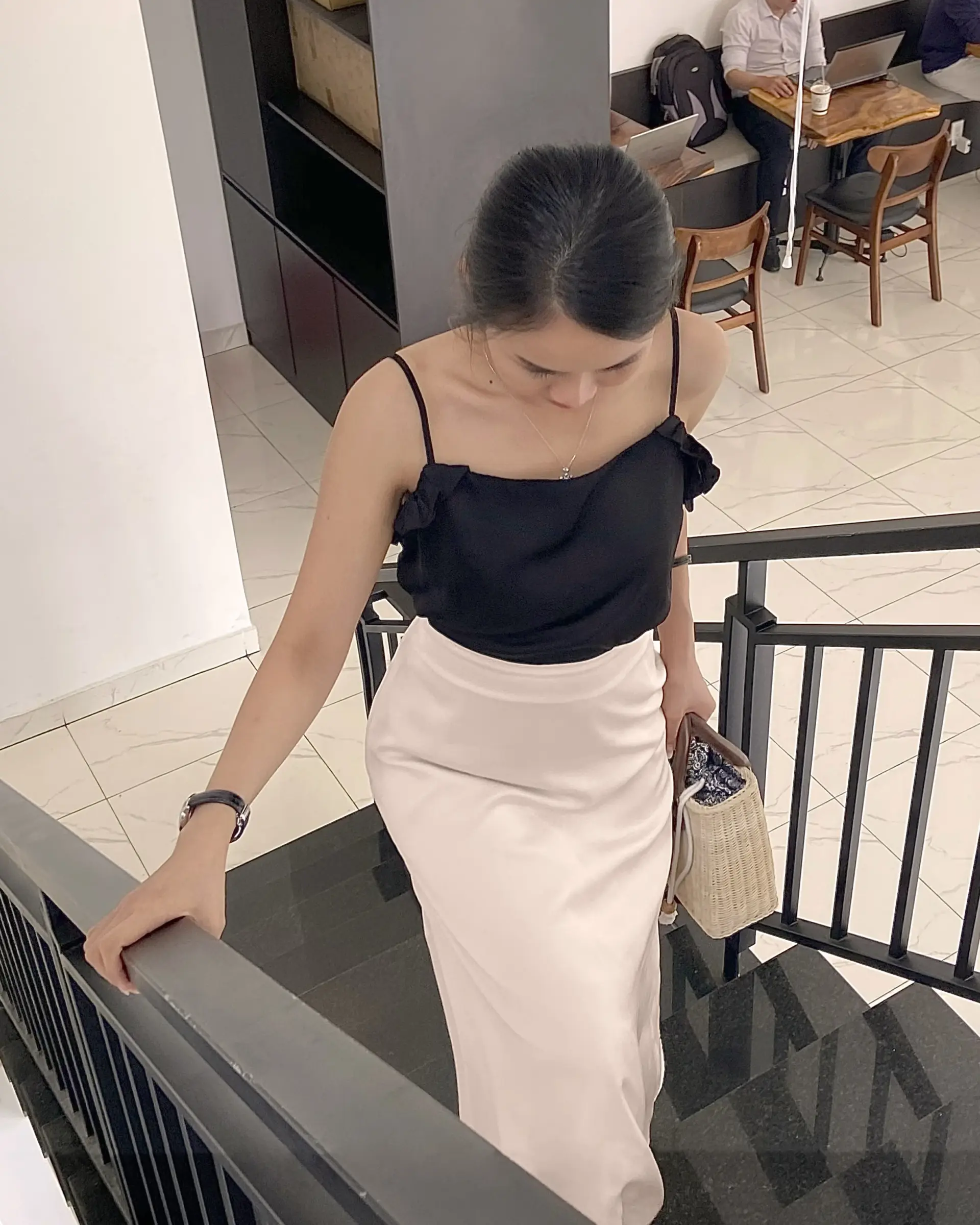 Woman in black top descends staircase, holding woven bag, captured in minimalist indoor setting.
