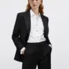 Woman in a black suit with a white shirt, hands in pockets, posing confidently against a plain background.
