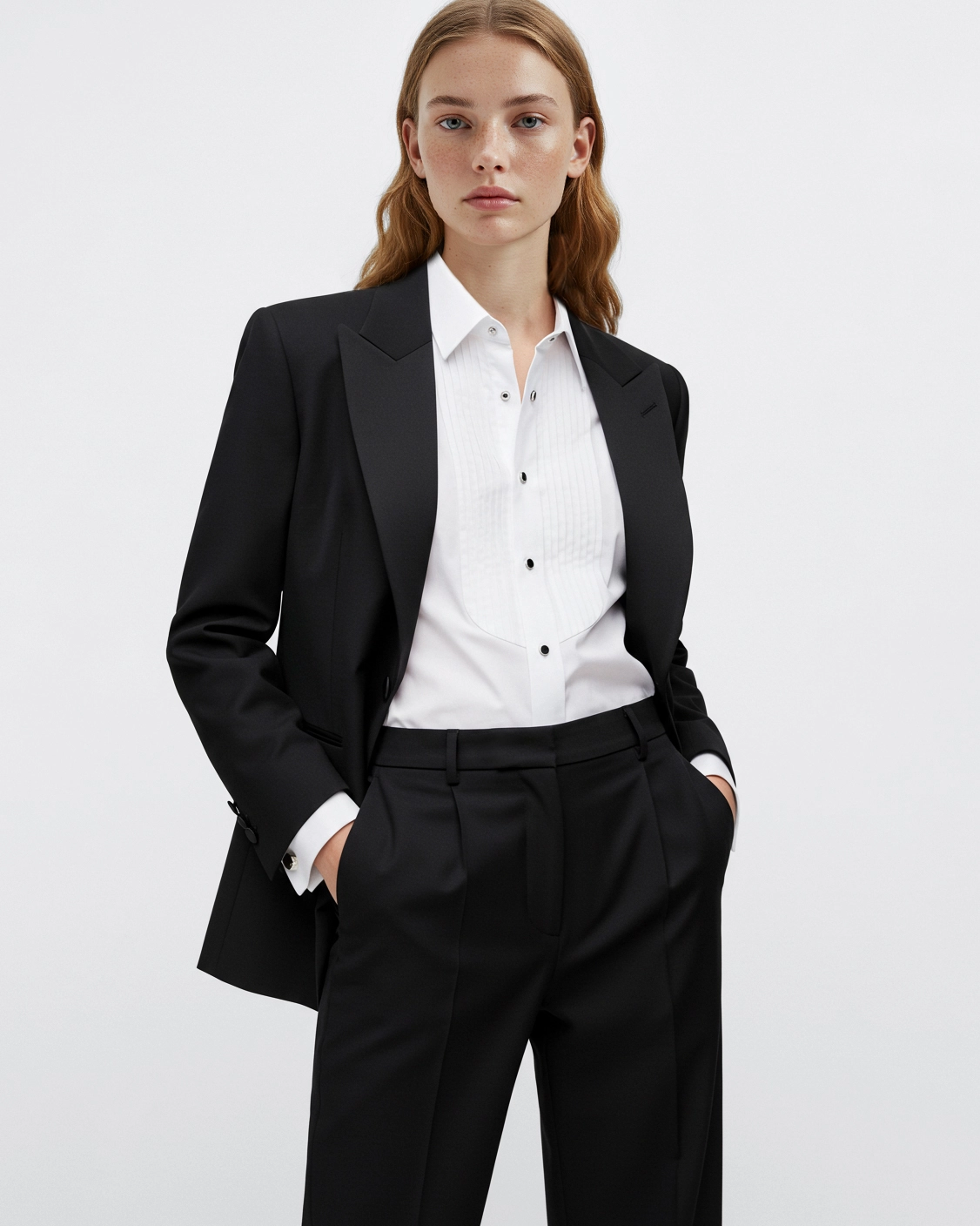 Woman in a black suit with a white shirt, hands in pockets, posing confidently against a plain background.