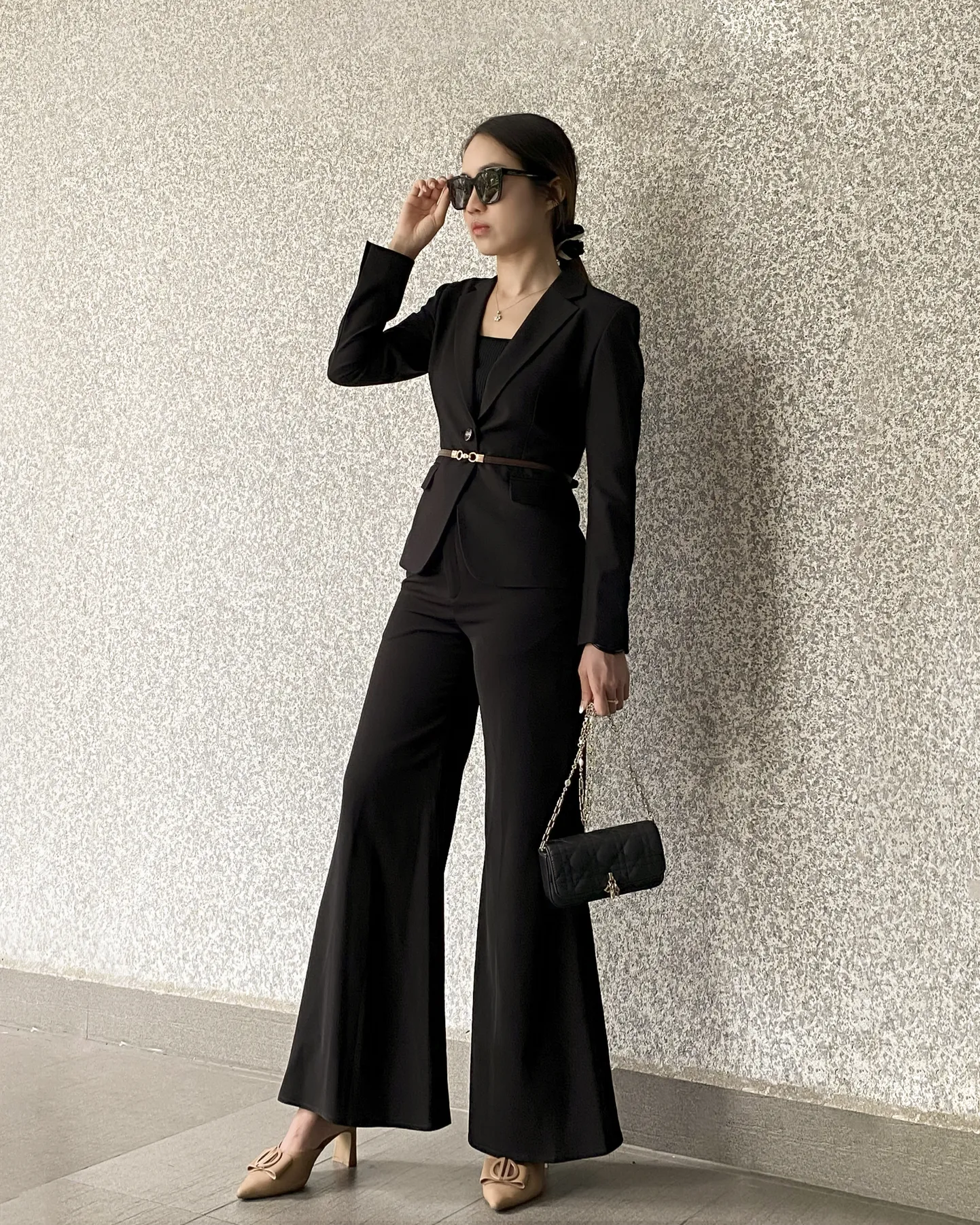 Woman in stylish tailored black trousers suit and sunglasses holds a handbag, leaning against a textured wall.