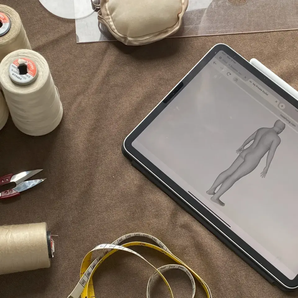 Fabric design tools, including tablet with 3D model, thread spools, measuring tape, and snips on a brown surface.