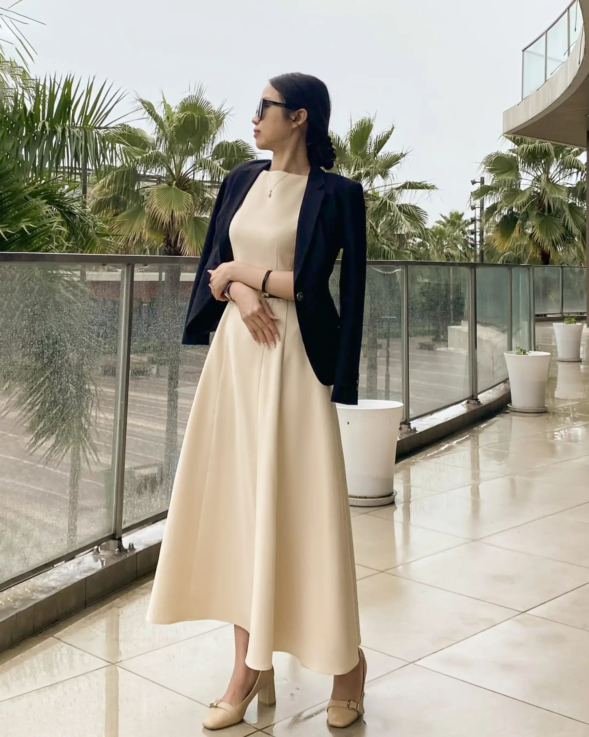 Elegant woman in beige tailored flare dress and black tailored blazer on balcony with palm trees in background, looking stylishly to the side.
