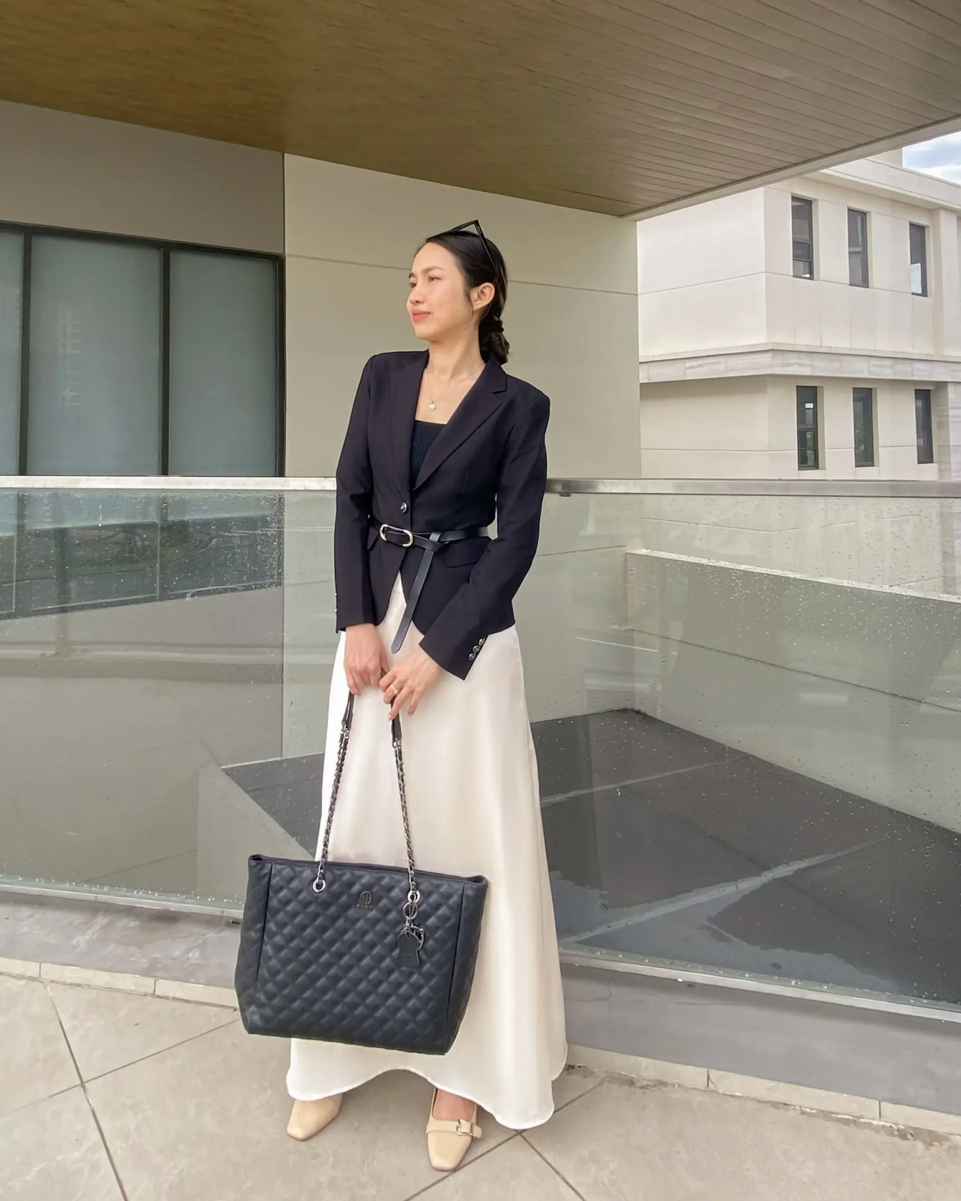 Woman in stylish black tailored blazer and white tailored silk skirt holding a quilted handbag on a modern building balcony.