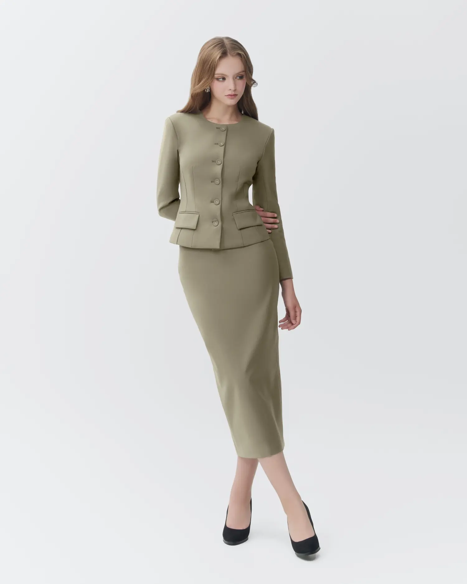 Woman in olive skirt suit with long sleeves and button detail, standing gracefully in black heels.