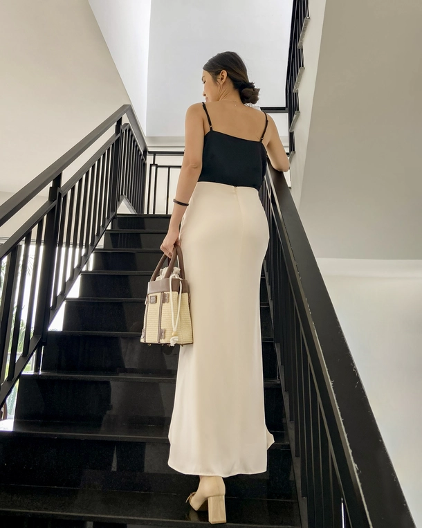 Woman in a stylish tailored silk outfit walking upstairs, carrying a raffia woven handbag, showcasing modern fashion, indoors.