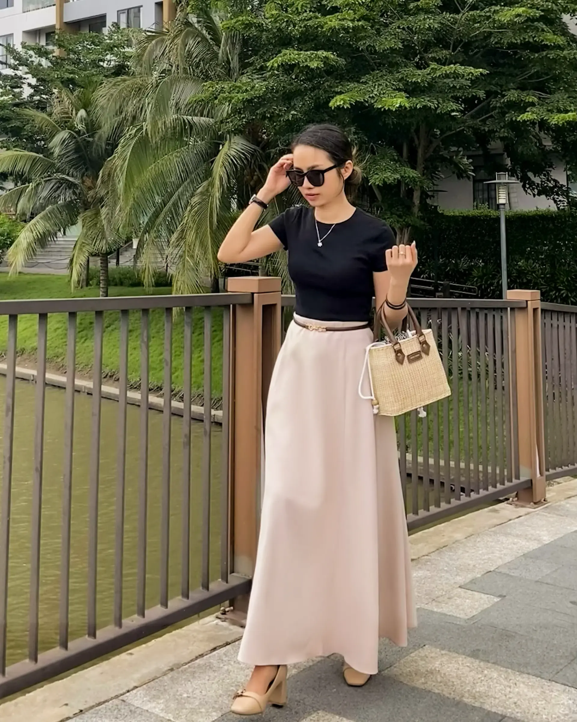 Woman in black top and tailored circle skirt, holding a handcrafted rattan handbag, standing by a riverside railing with tropical trees in the background.