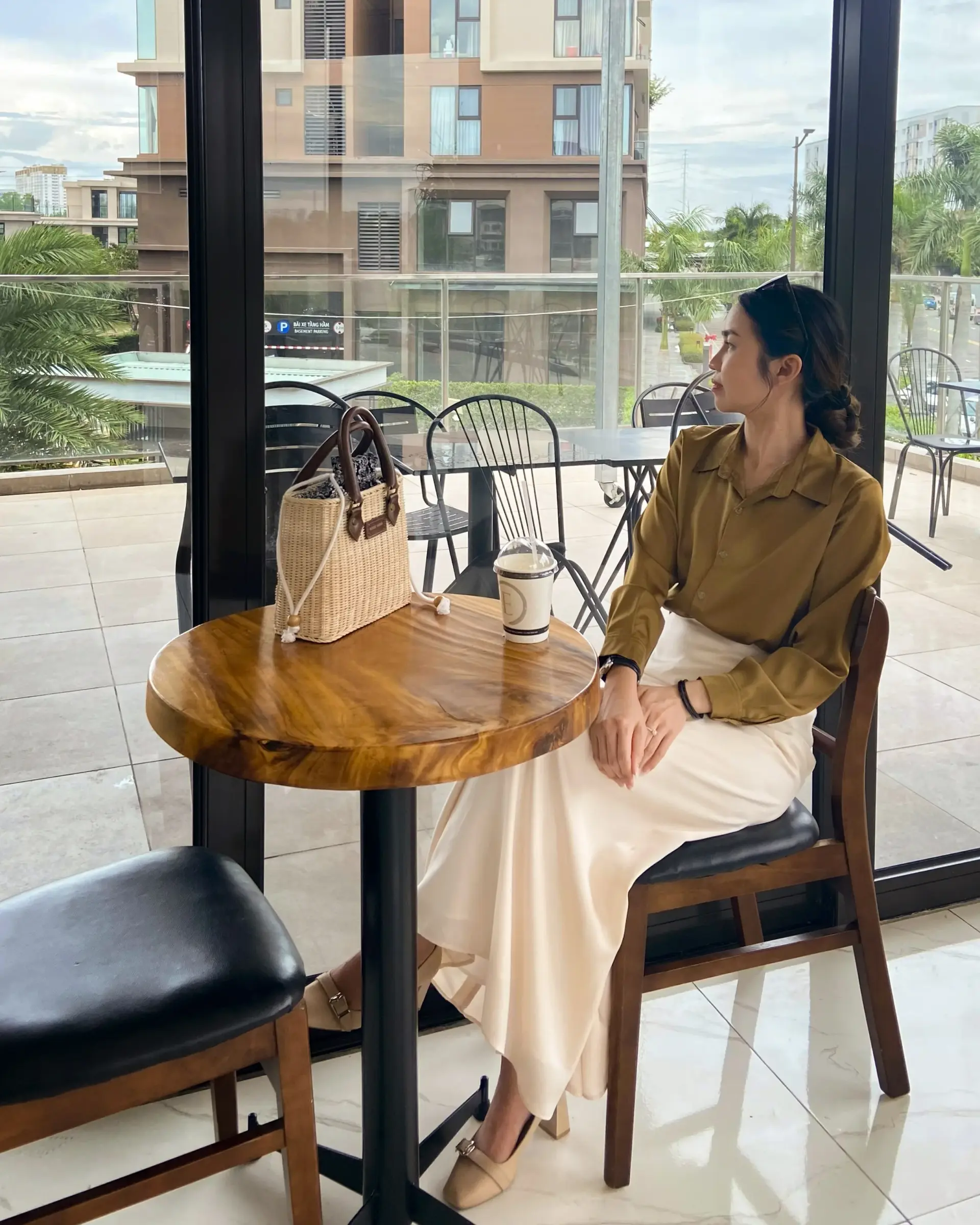 Woman in a tailored silk shirt and tailored silk skirt sitting at a café, gazing outside thoughtfully with a rattan handbag on the table.