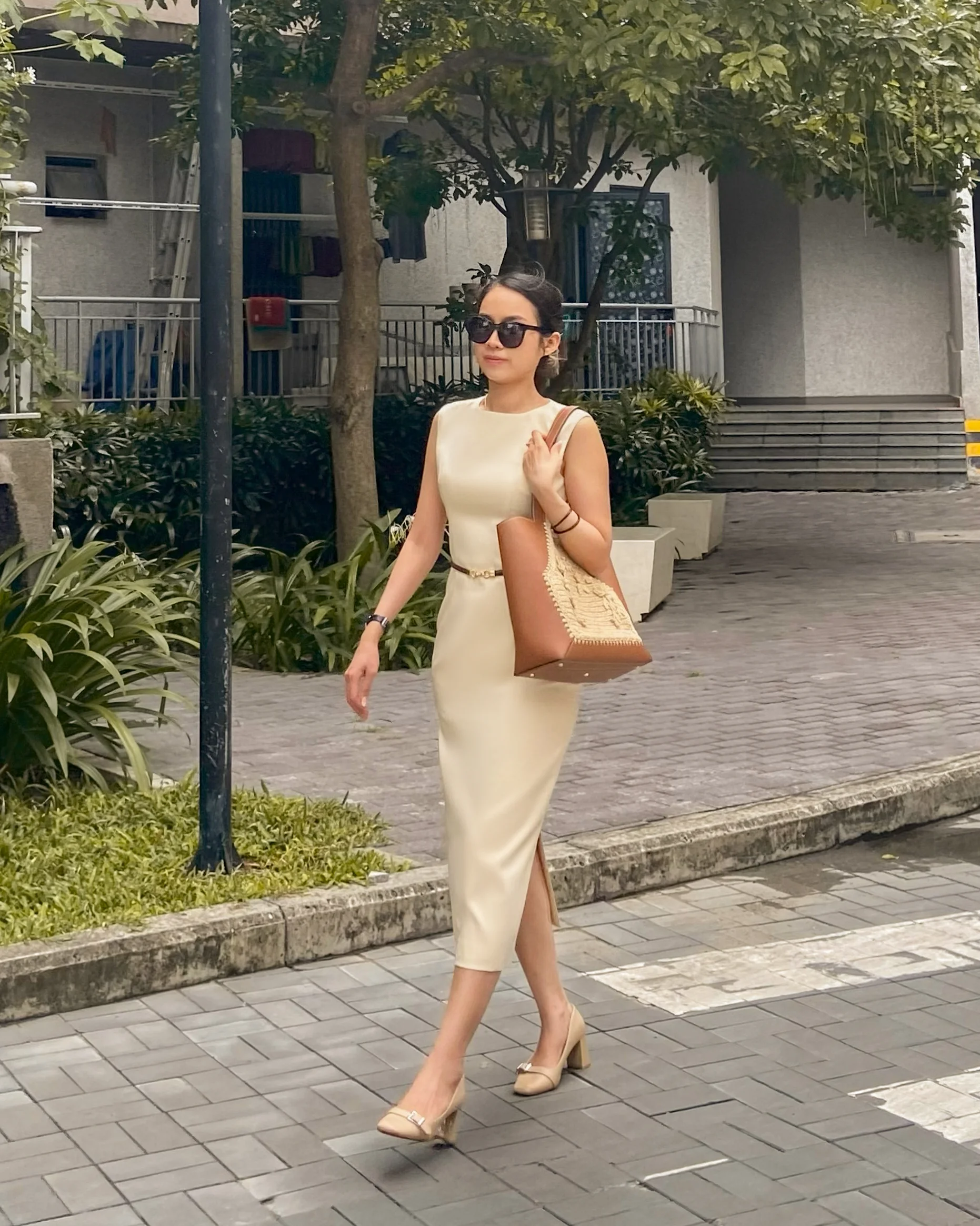 Woman in beige tailored sheath dress with sunglasses and brown crocheted raffia bag walking on sidewalk.