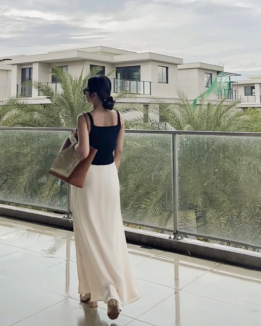 Woman in elegant tailored silk skirt with crocheted raffia bag walking on terrace with modern buildings and palm trees in the background.