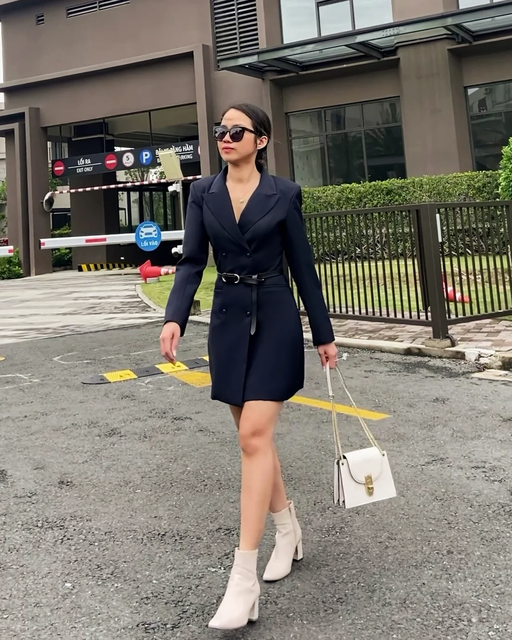Fashionable woman in a navy tailored blazer dress and boots walking confidently, carrying a white handbag outdoors.