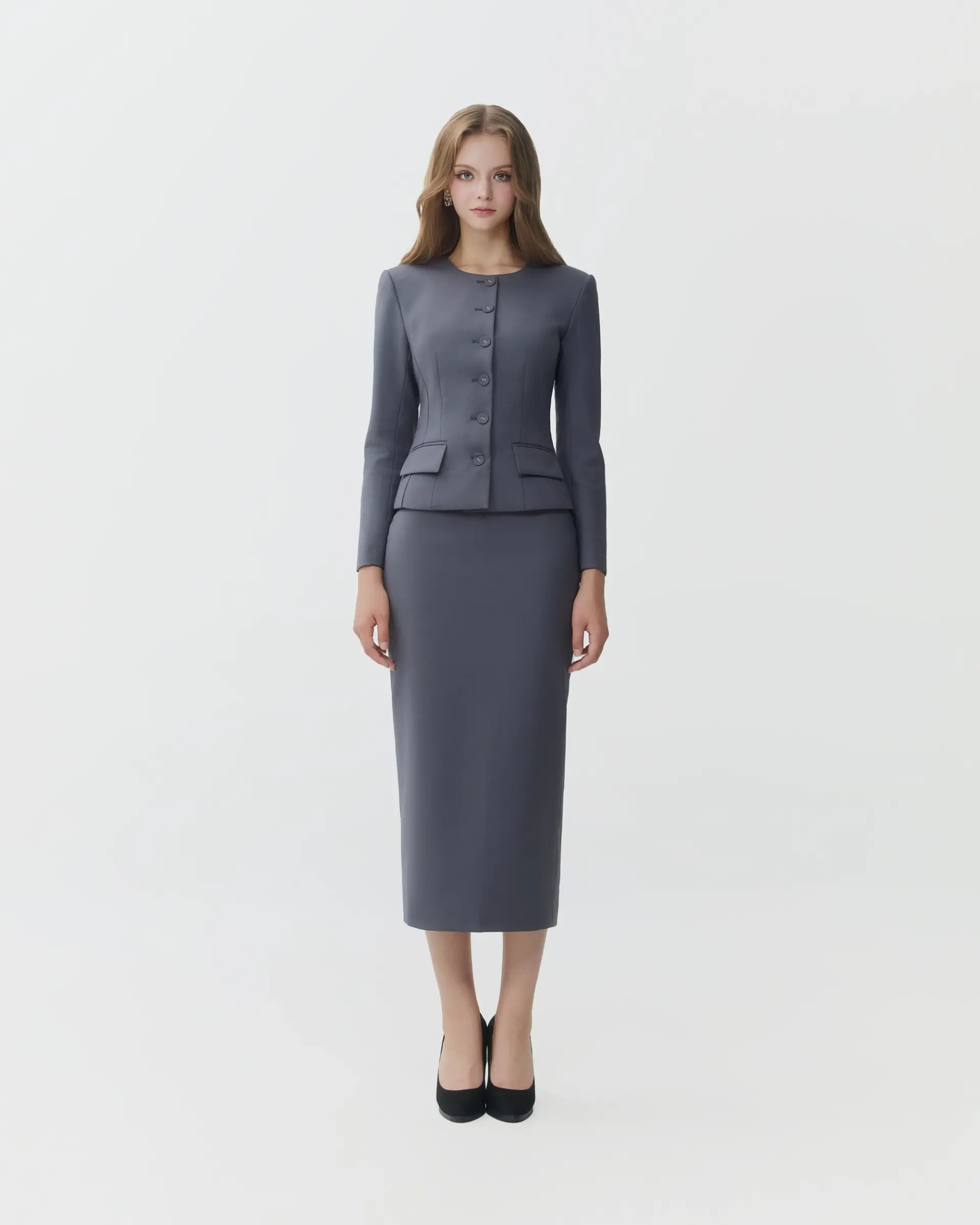 Woman in formal gray suit with long skirt and black heels, standing against a white background.